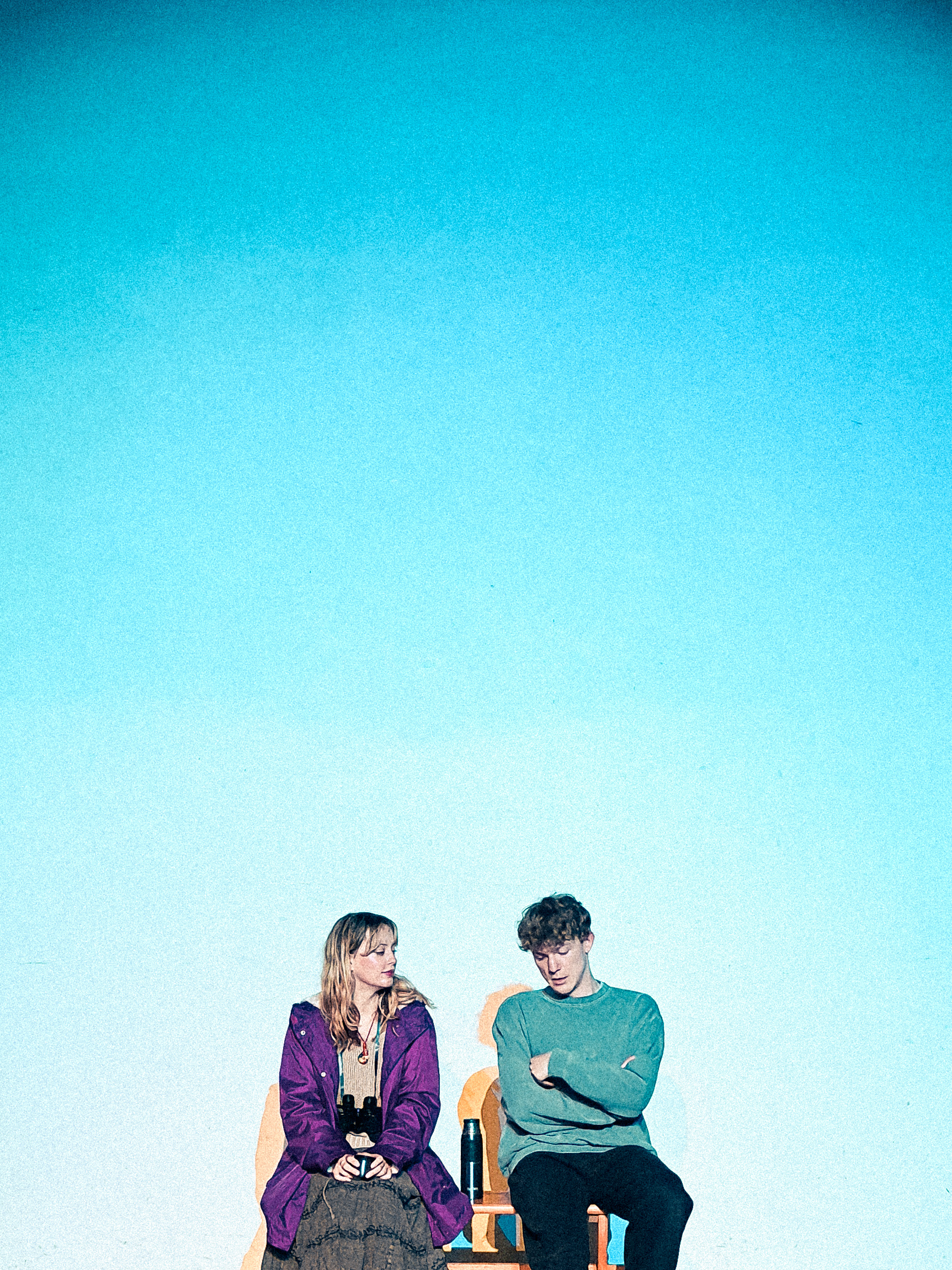 Tech still of actors Andy and Gwithian sitting side by side on a bench, against a blue background. 