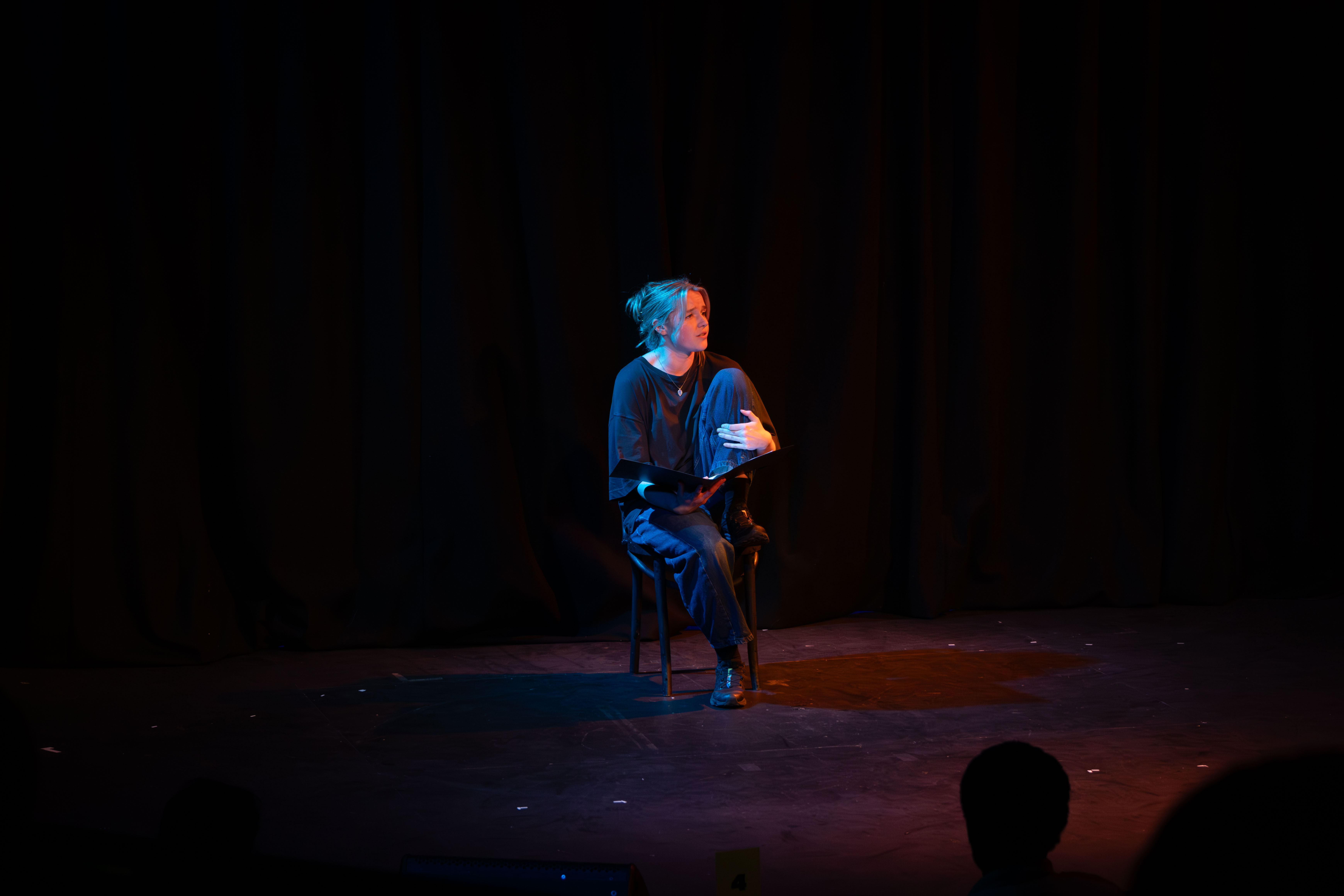 Liv onstage as Mag, seated. Teal and orange light.