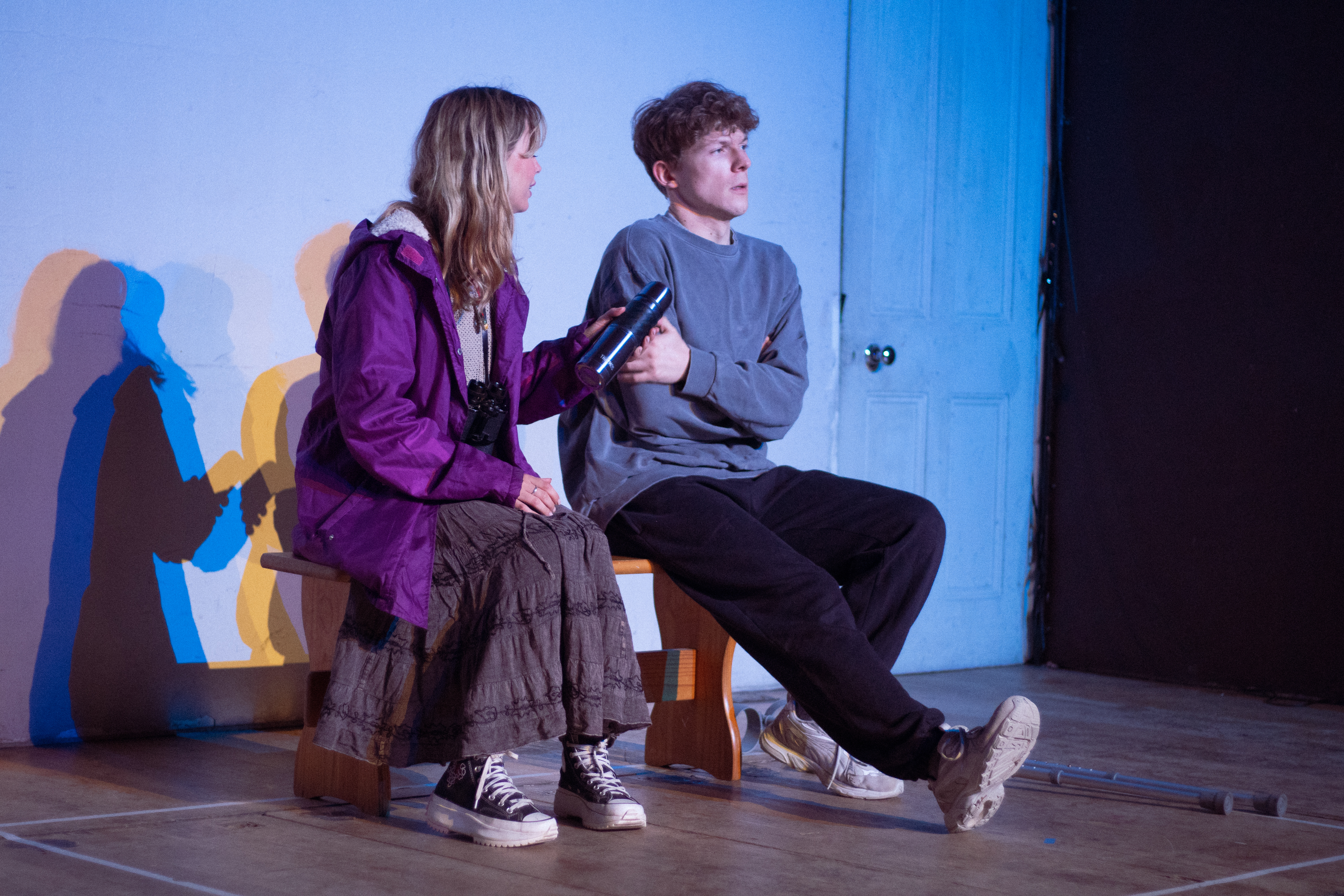 Tech still of actors Andy and Gwithian sitting side by side on a bench. Andy is holding out a flask of hot chocolate. 