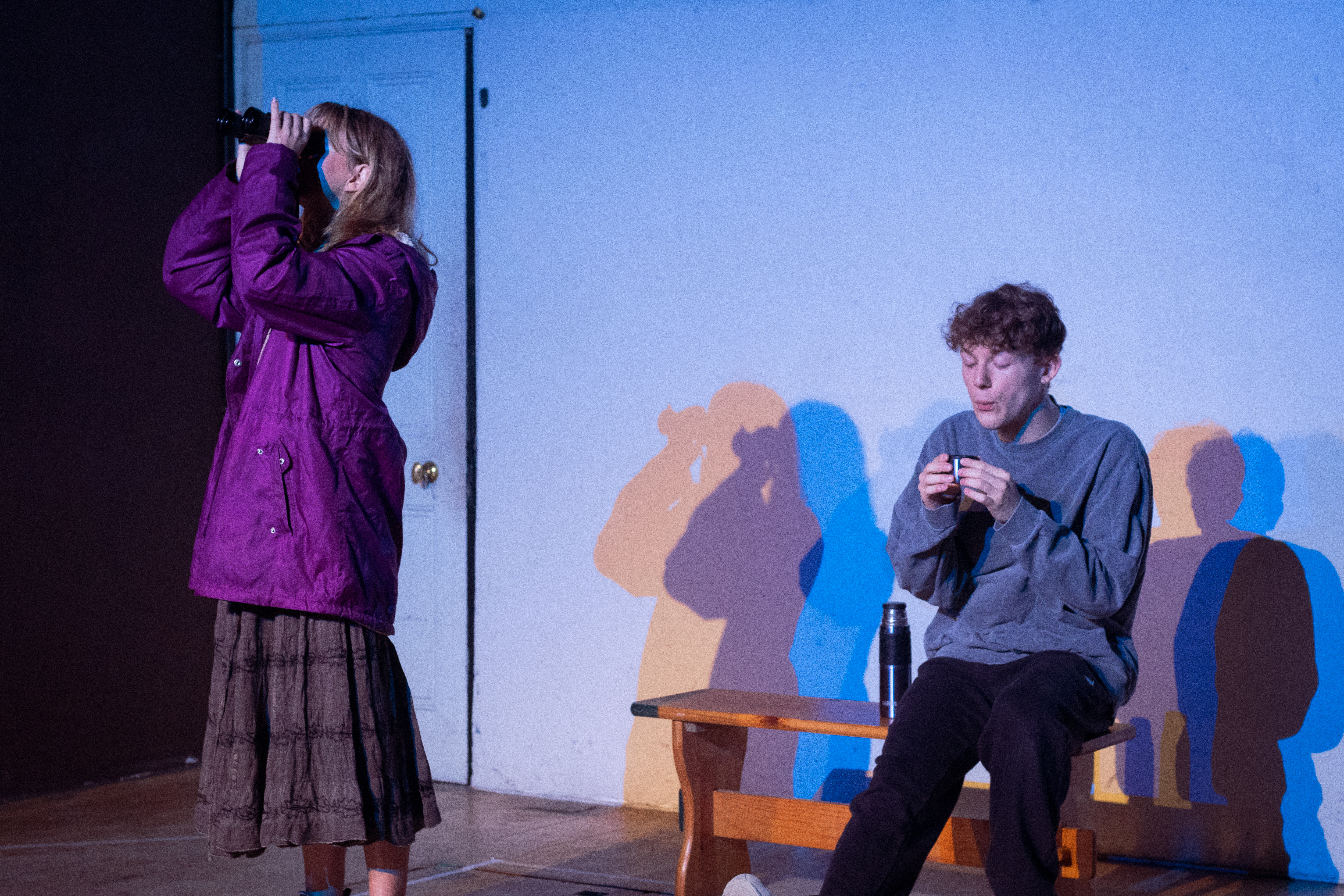 Tech still of actors Andy and Gwithian. Andy is looking out front with a pair of binoculars. Gwithian is sitting on a bench behind her, blowing on a cup of hot chocolate.