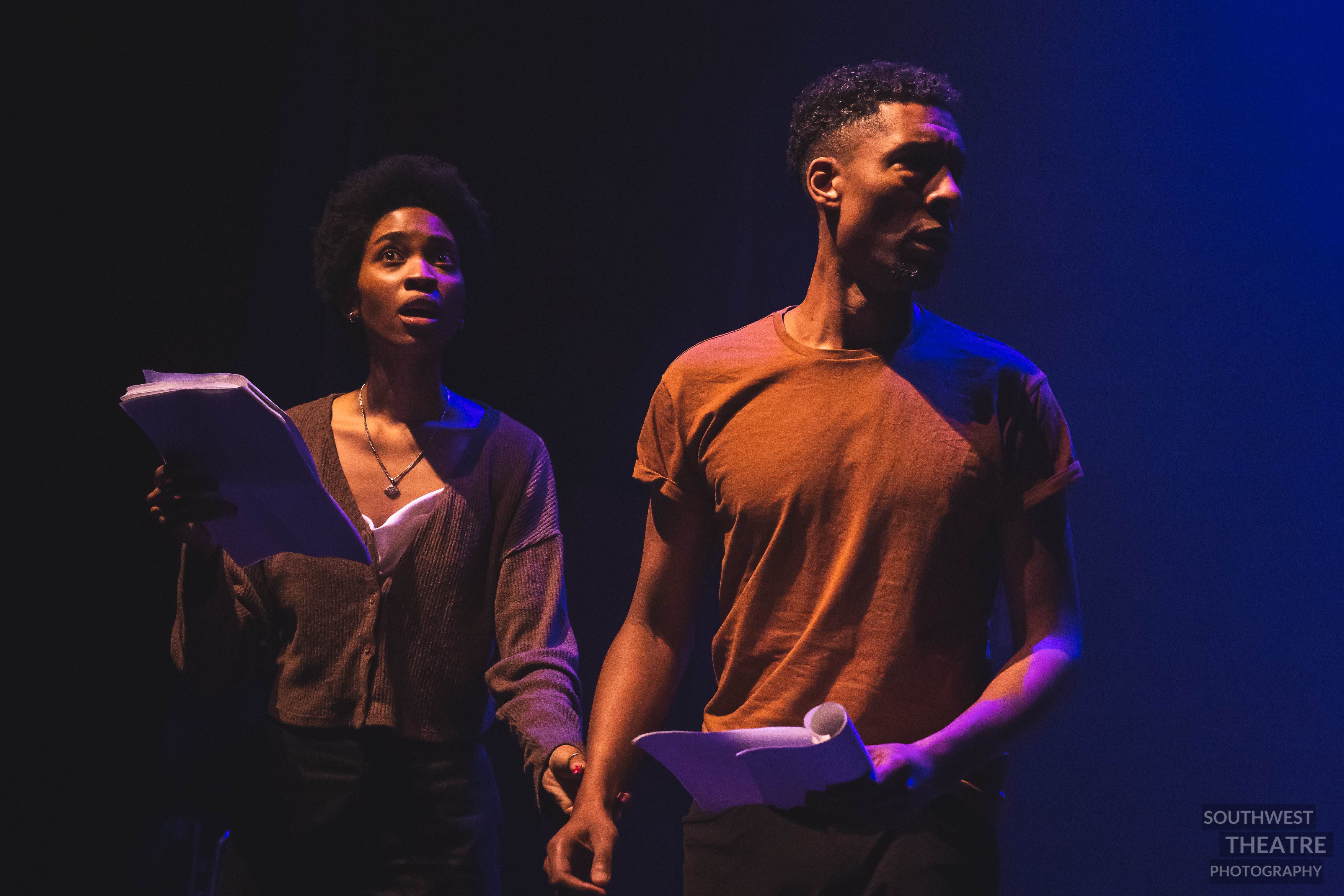 N’Dea, playing a centipede, and Emile, playing Moth B, are standing close together on stage. N’Dea is looking upwards and Emile is looking away. N’Dea is holding Emile’s wrist.