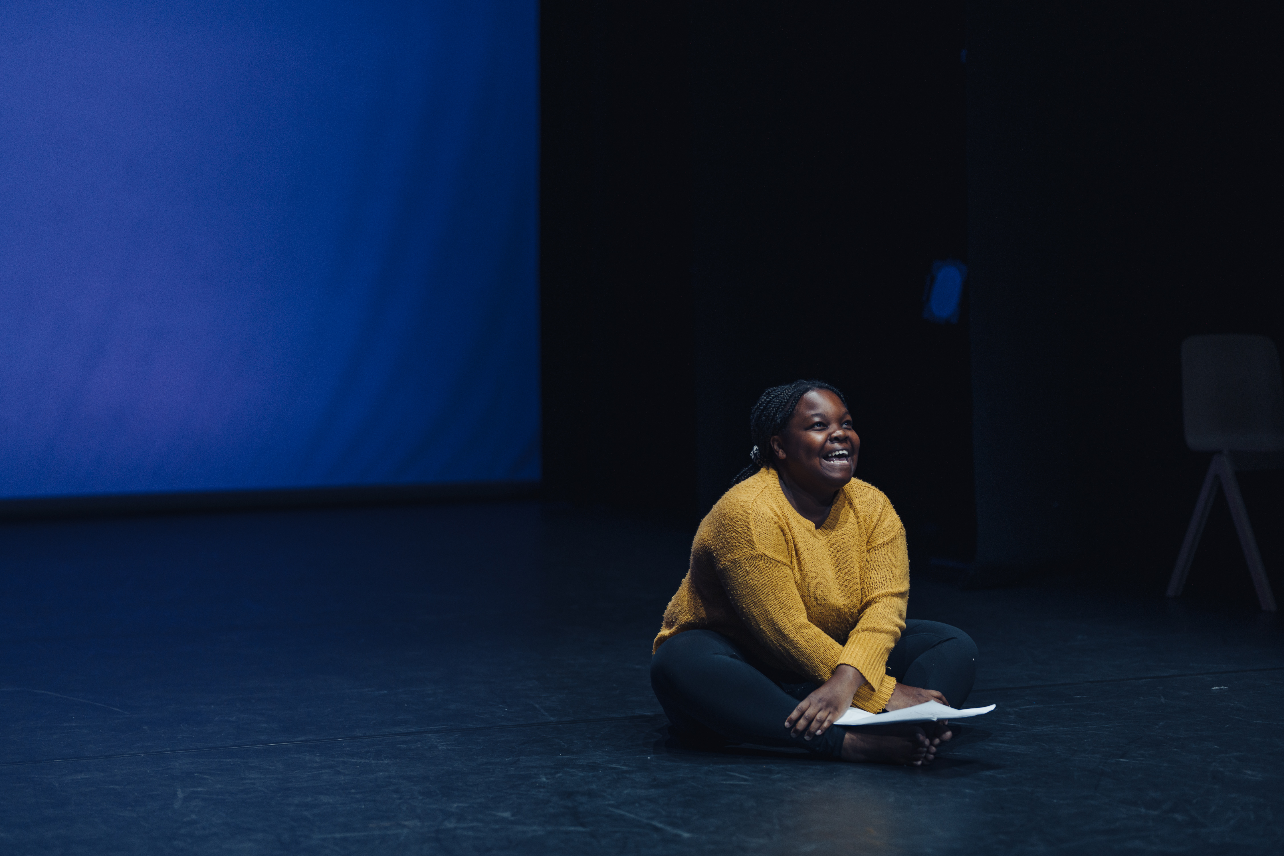 Corinne, a black woman wearing a yellow jumper, is sitting cross-legged on the floor of the stage. She is smiling and looking up, as if towards the sky.